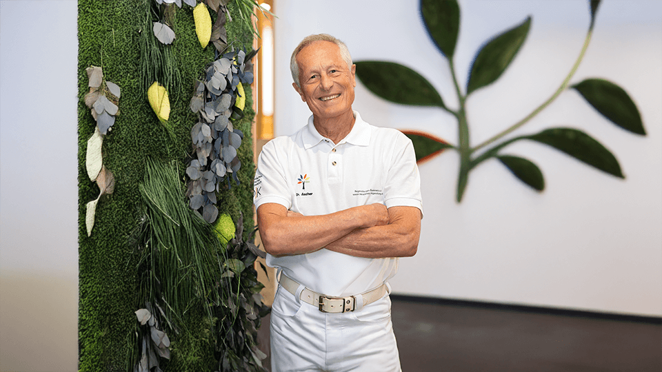 Dr. Gerhard Ascher steht vor einer grünen Pflanzenwand mit verschränkten Armen und lächelt
