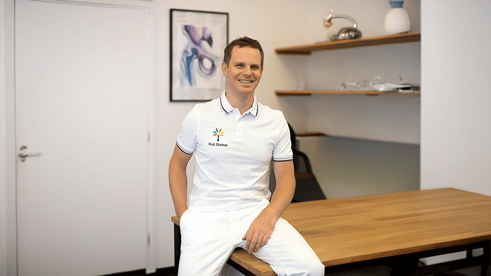 Porträt von Prof. Dr.Felix Greimel – Facharzt für Orthopädie sitzend auf einem Holztisch in einem Büro