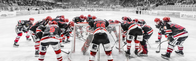 Eishockeyspieler in schwarzer und roter Ausrüstung beugen sich vor dem Tor in einem Stadion zusammen.