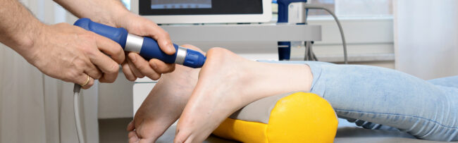 Hand hält ein blaues medizinisches Gerät zur Stoßwellentherapie an die Ferse eines liegenden Beins, das auf einem gelben Kissen ruht.